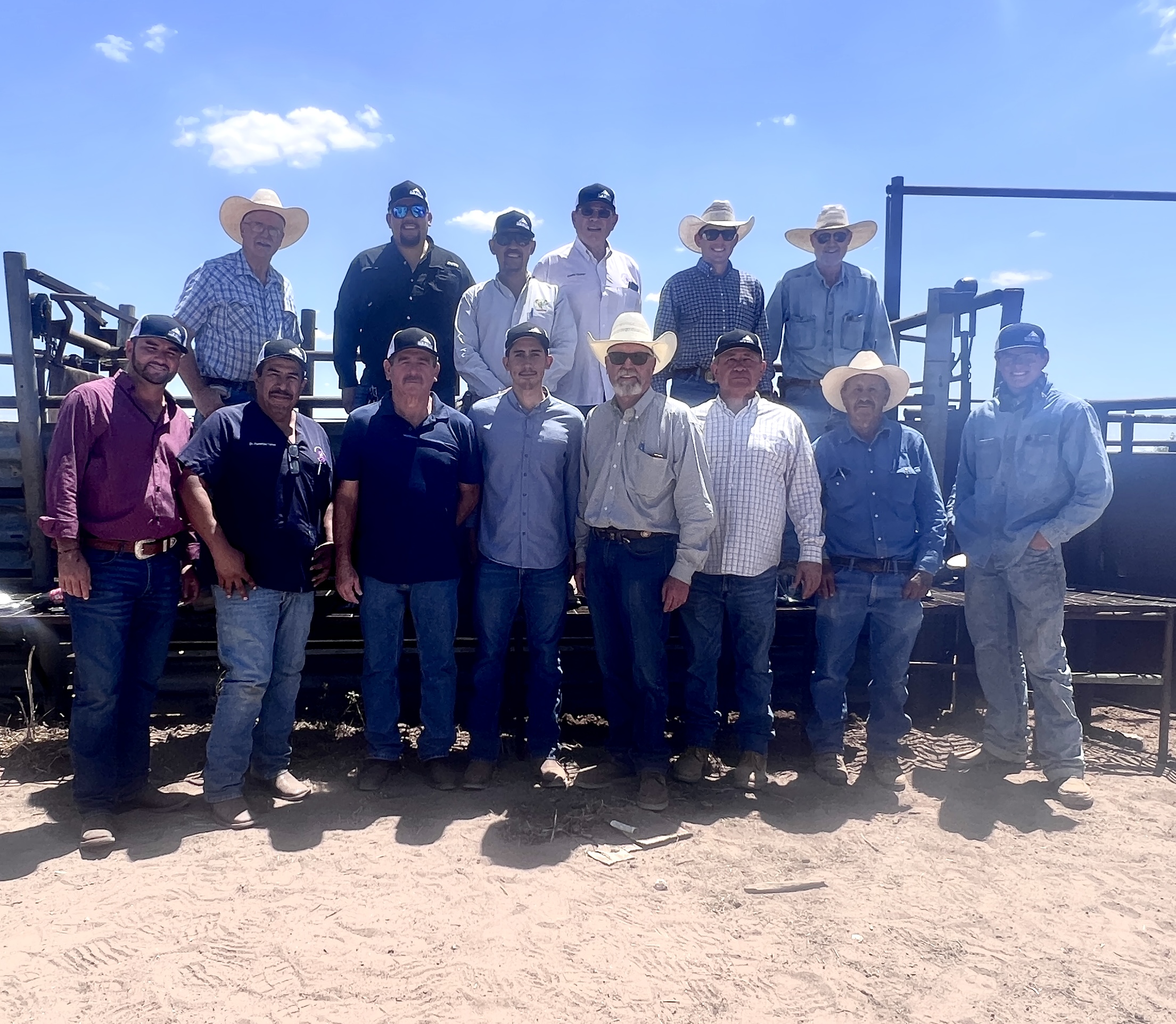 Fourteen men stand in a group posing for a photo outside a cattle pen.