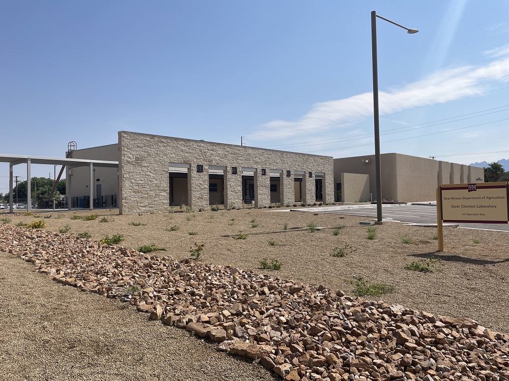 Paisaje abierto con rocas grandes y pequeñas de color canela que conducen a una pared de roca y un pasillo y un letrero para el Laboratorio Estatal de Química del Departamento de Agricultura de Nuevo México.