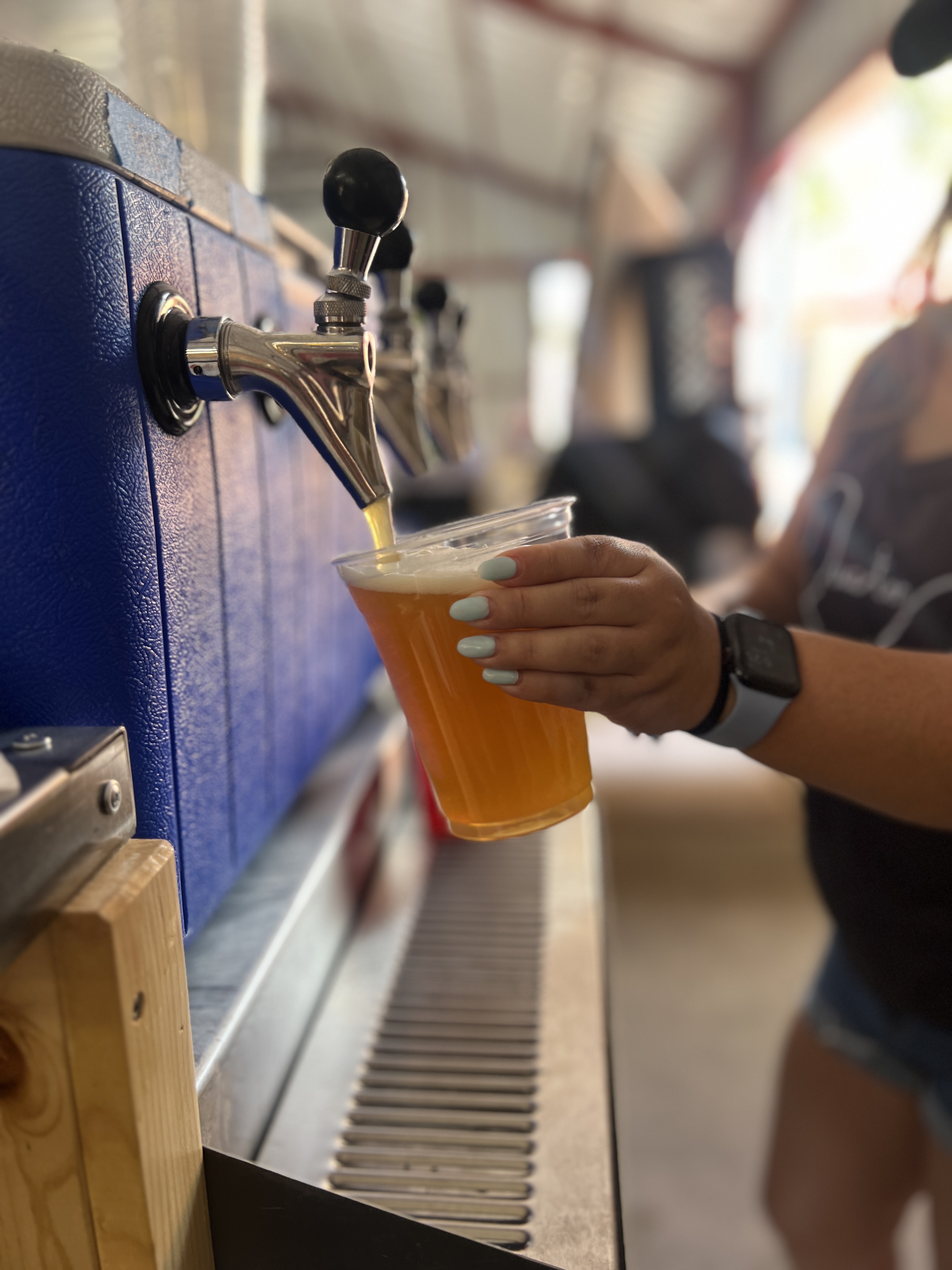 Close-up of a beer tap pouring golden draft beer into a clear plastic cup held by someone wearing a smartwatch.