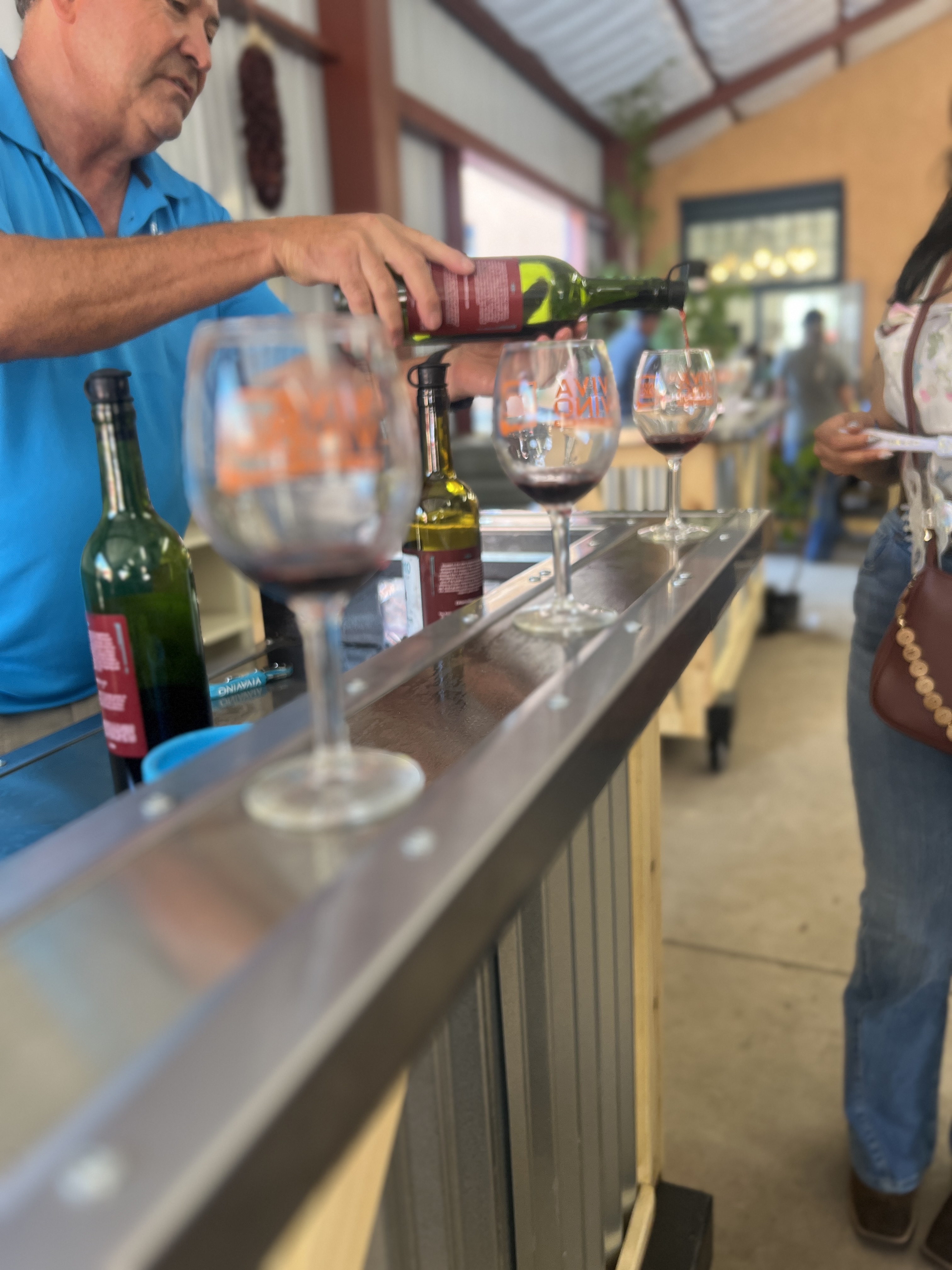 Person pouring red wine from a bottle into tasting glasses lined up on a bar counter at a winery tasting room.