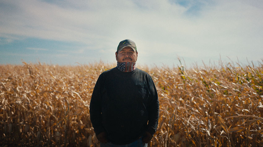 A man wearing a baseball cap and a neck gator stands in the middle of a corn field 