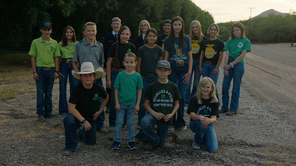 A group of 12 students dressed in 4-H and FFA T-shirts stand and kneel together smiling toward the camera, with an orchard in the background.