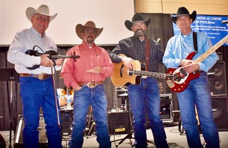 four men in cowboy hats pose on stage in front of a drum set and large speakers. Each person is holding a musical instrument. 