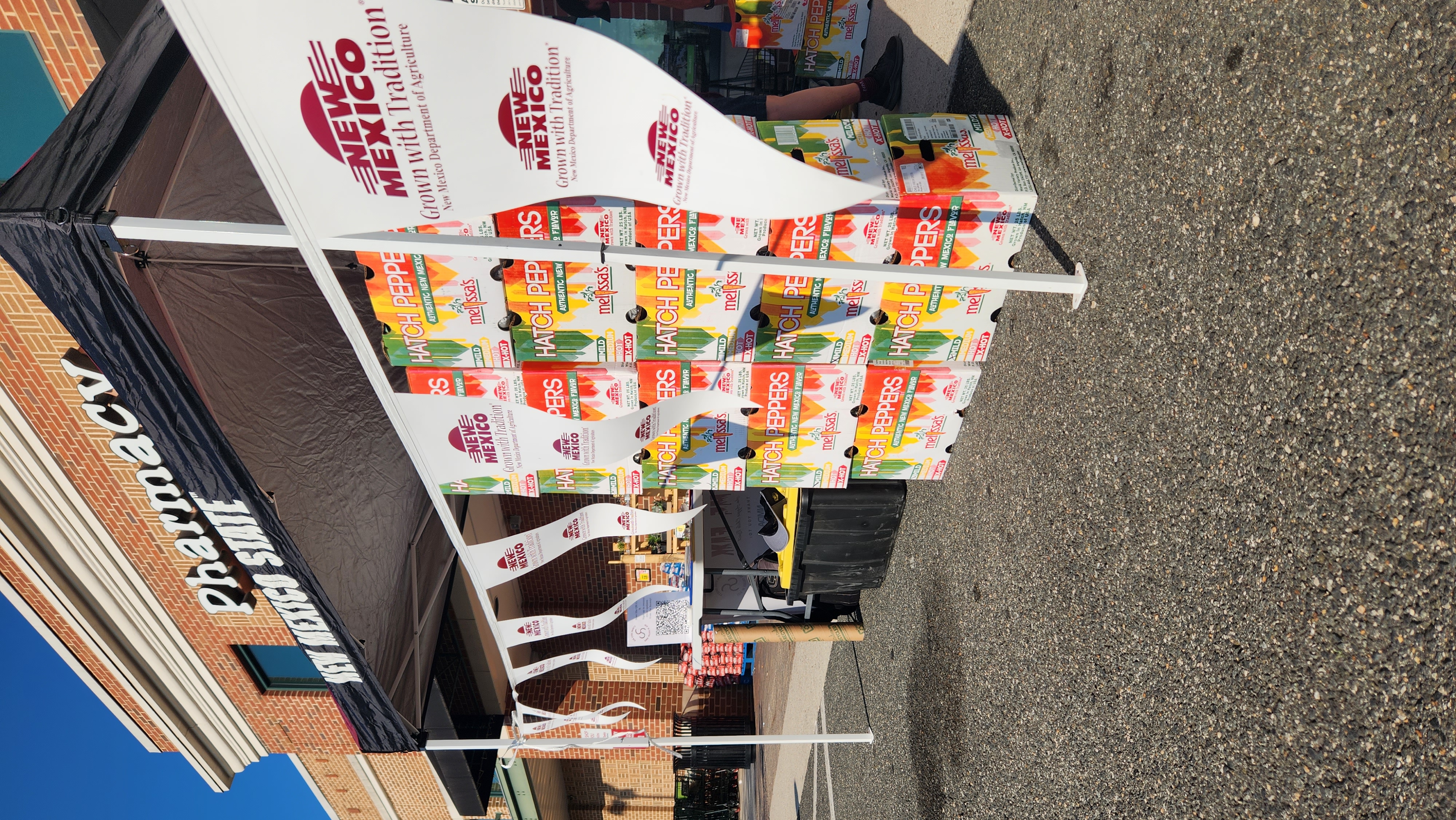 Boxes of Hatch green chile stacked under a canopy with New Mexico Department of Agriculture flags at a Harris Teeter grocery store in Alexandria, Virginia.