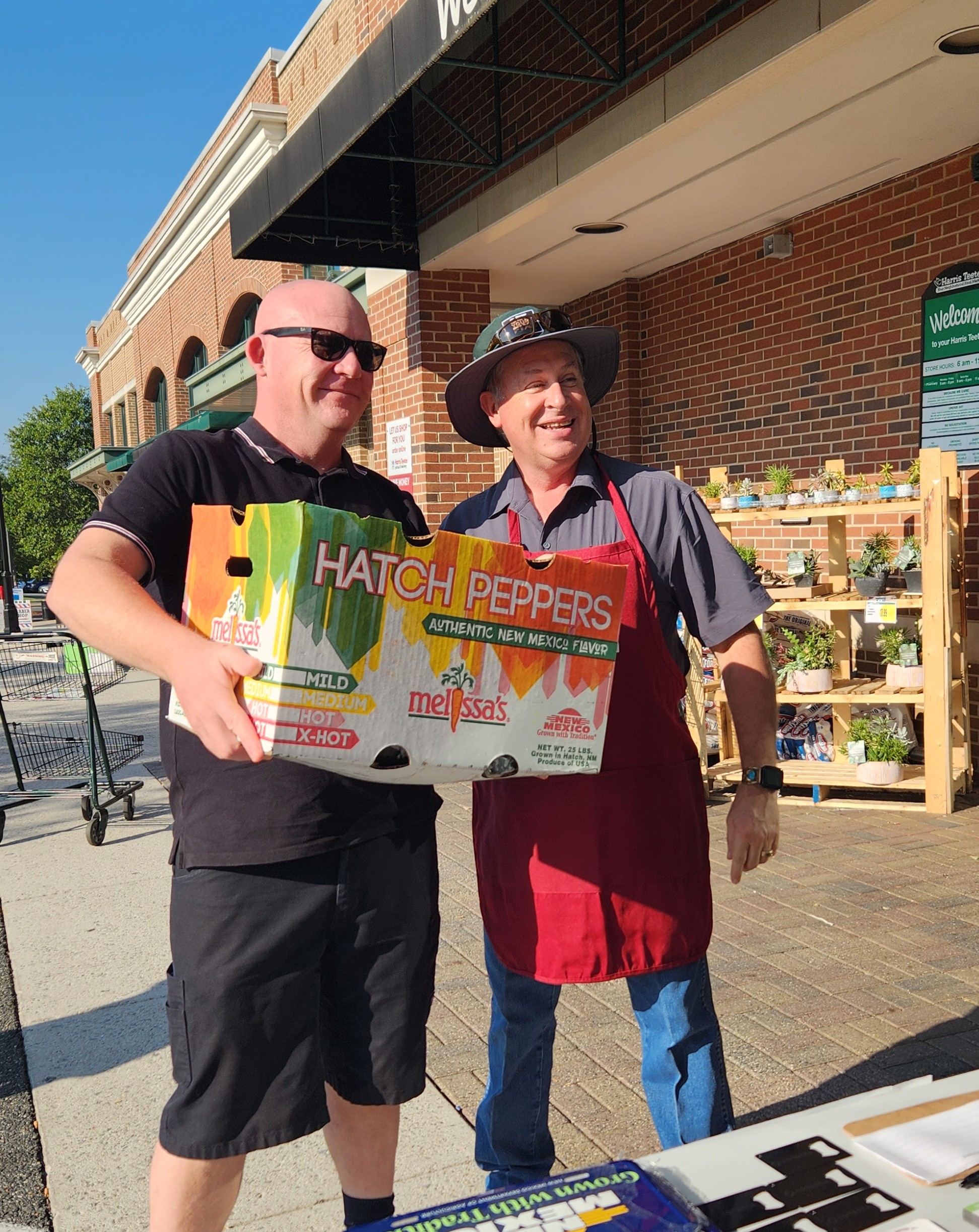 Two men smiling while holding a box labeled “Hatch Peppers – Authentic New Mexico Flavor” outside a grocery store during a chile roast event. 