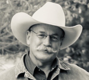 A black and white image of a man with a white cowboy hat and thin glasses hiding a smile through his white mustache with blurred forestry in the background. 