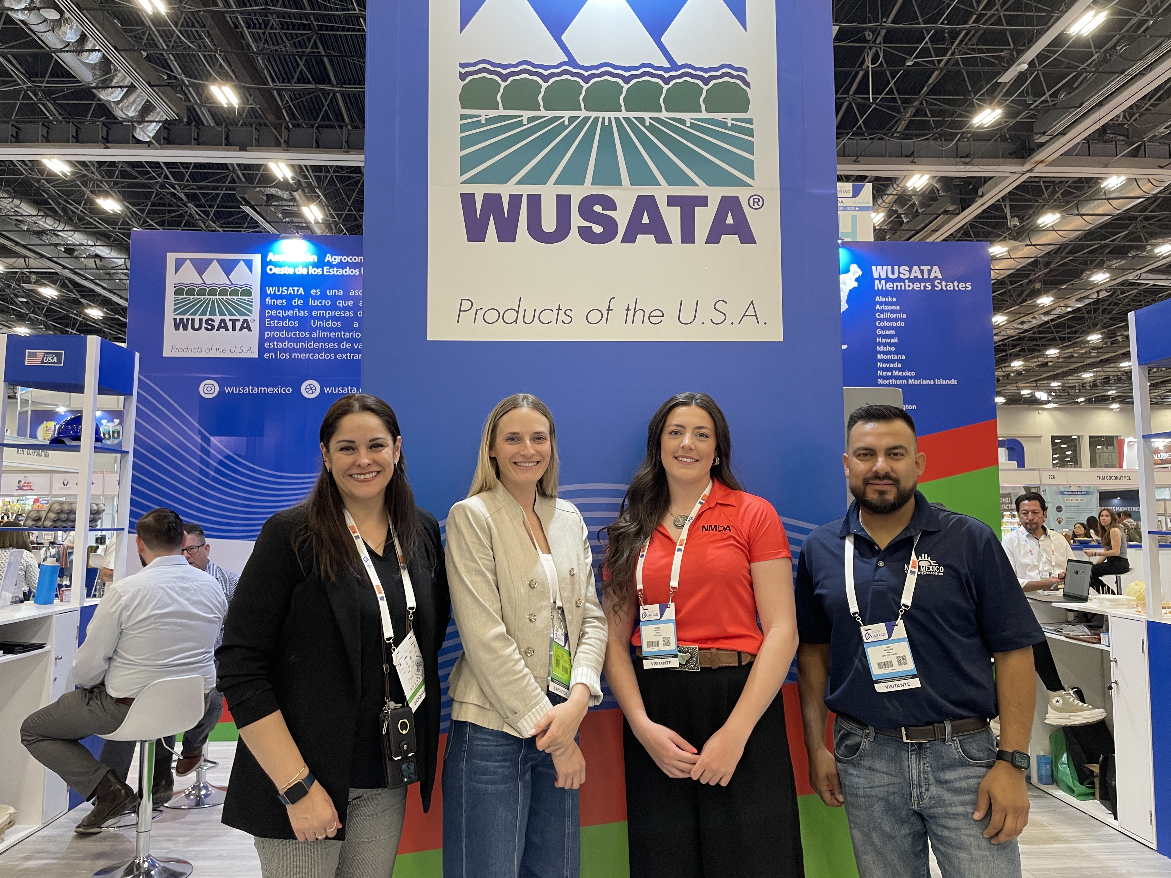 Four people stand in front of a trade show booth displaying the “Products of the U.S.A.” logo at an indoor exhibition hall.