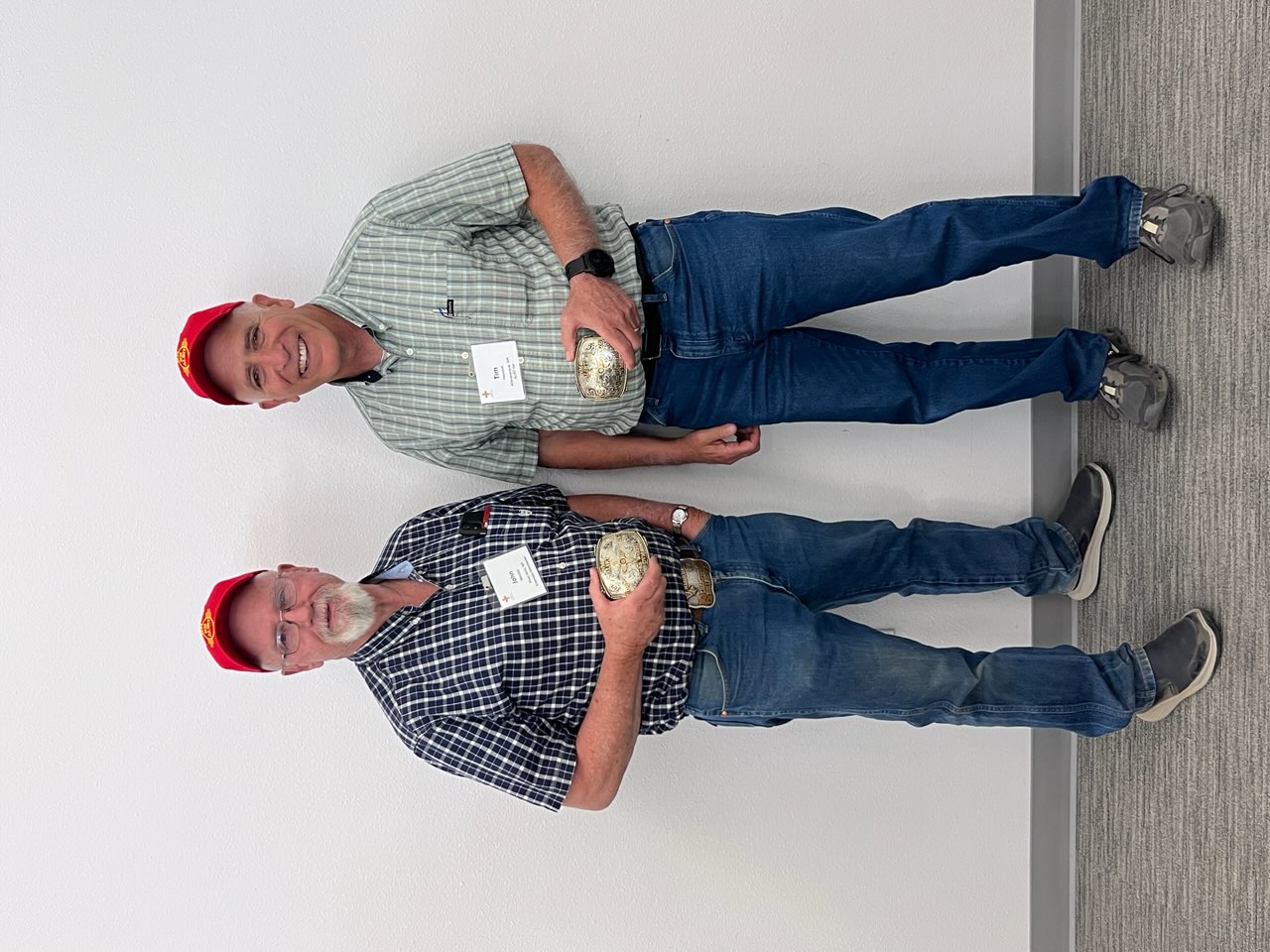 Two men stand side by side against a white wall, both wearing plaid shirts and red hats while holding their Agriculture Legacy Awards.