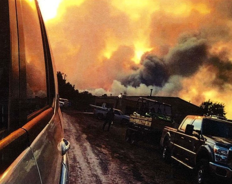 The view from a vehicle driving away from a building while a wildlife burns trees in the background. 