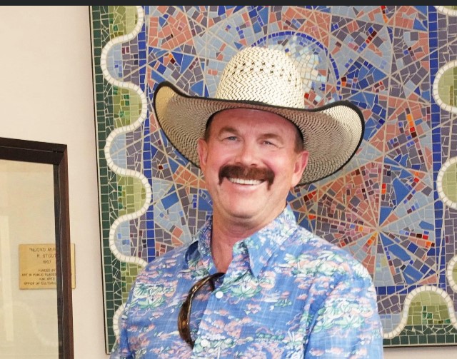 a man in a straw cowboy hat with a handlebar mustache smiling in a pastel colored shirt