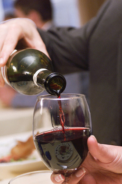 Red wine is poured out of a green wine bottle held by someone’s hand into a stemless, clear glass held by the person’s other hand.
