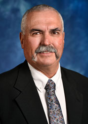 a man with short grey hair and grey mustache standing in front of a blue background wearing a black suit, white shirt and grey tie. 