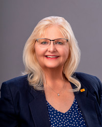 a blonde woman wearing glasses and an orange outfit standing in front of a white background. 