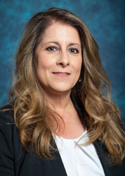 a woman with long, light brown hair wearing a white shirt and black jacket standing in front of a blue background. 