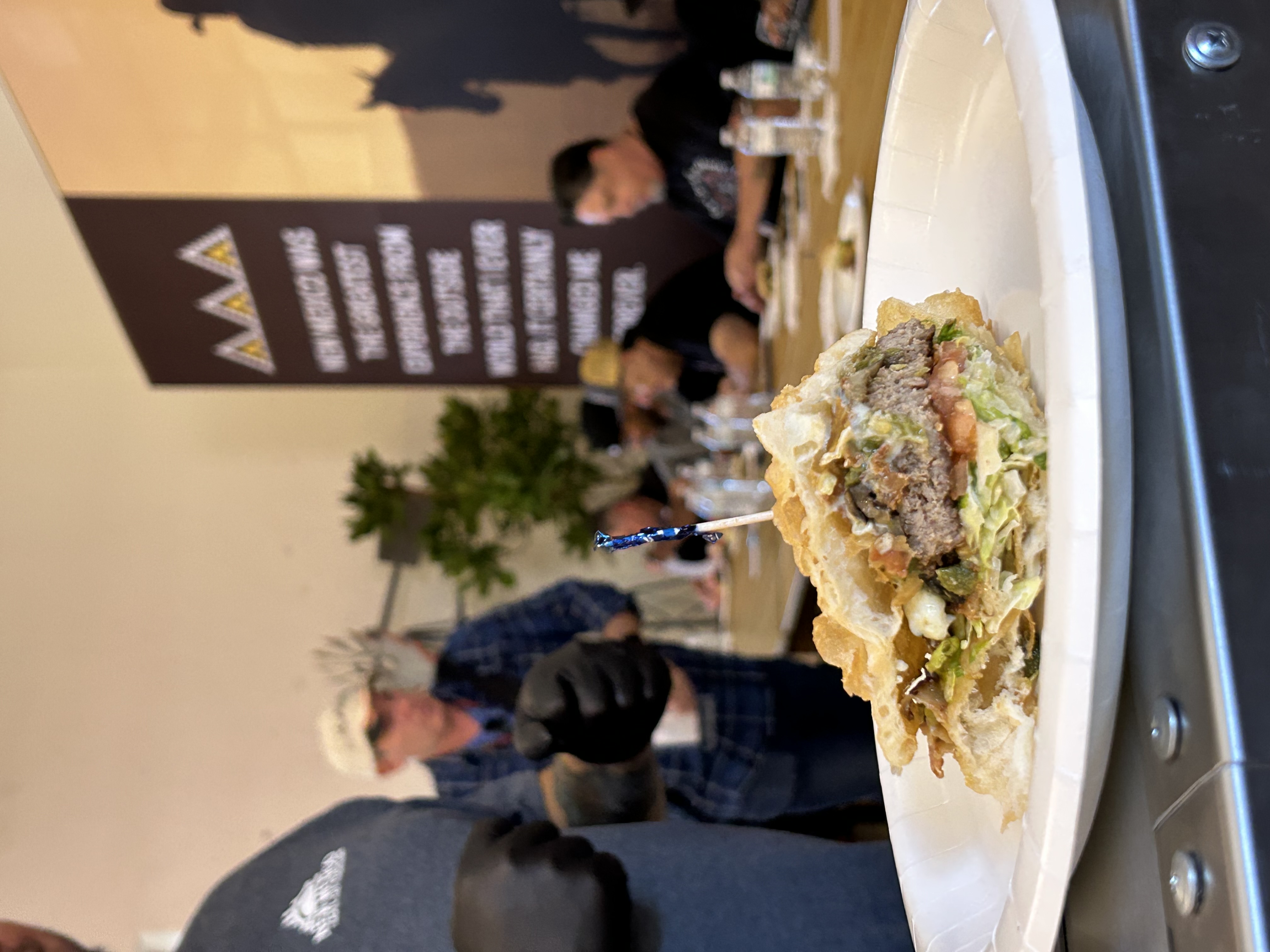 A green chile cheeseburger sits on a white plate, topped with green chile, cheese, lettuce, tomato and other toppings, with people seated at a judging table in the background.