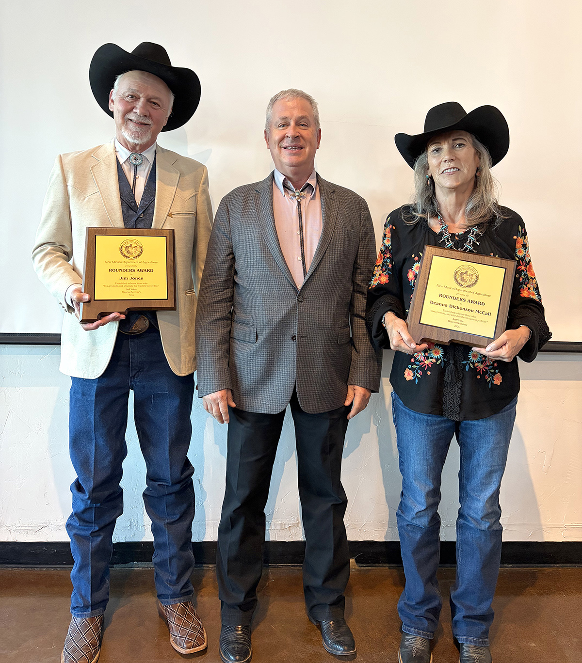 Three people pose indoors; two in cowboy hats hold award plaques while the person in the middle stands between them.