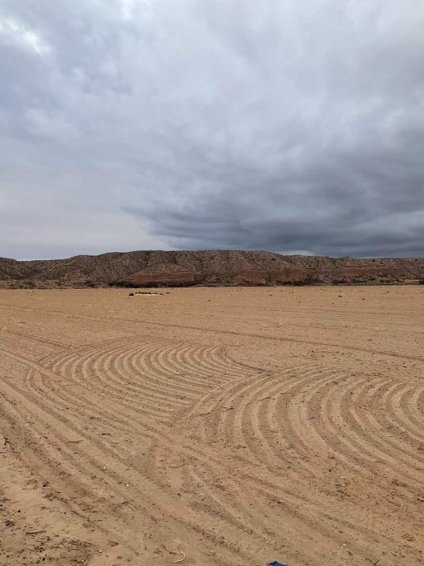 Una amplia y plana extensión de suelo desértico con huellas de neumáticos arqueadas en primer plano, que conduce hacia colinas bajas y escarpadas bajo un cielo dramático y nublado en el Valle Inferior del Río Grande.