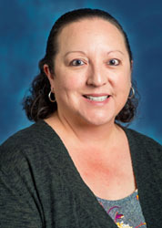 a woman with short black curly hair wearing a dark grey sweater standing in front of a blue background.